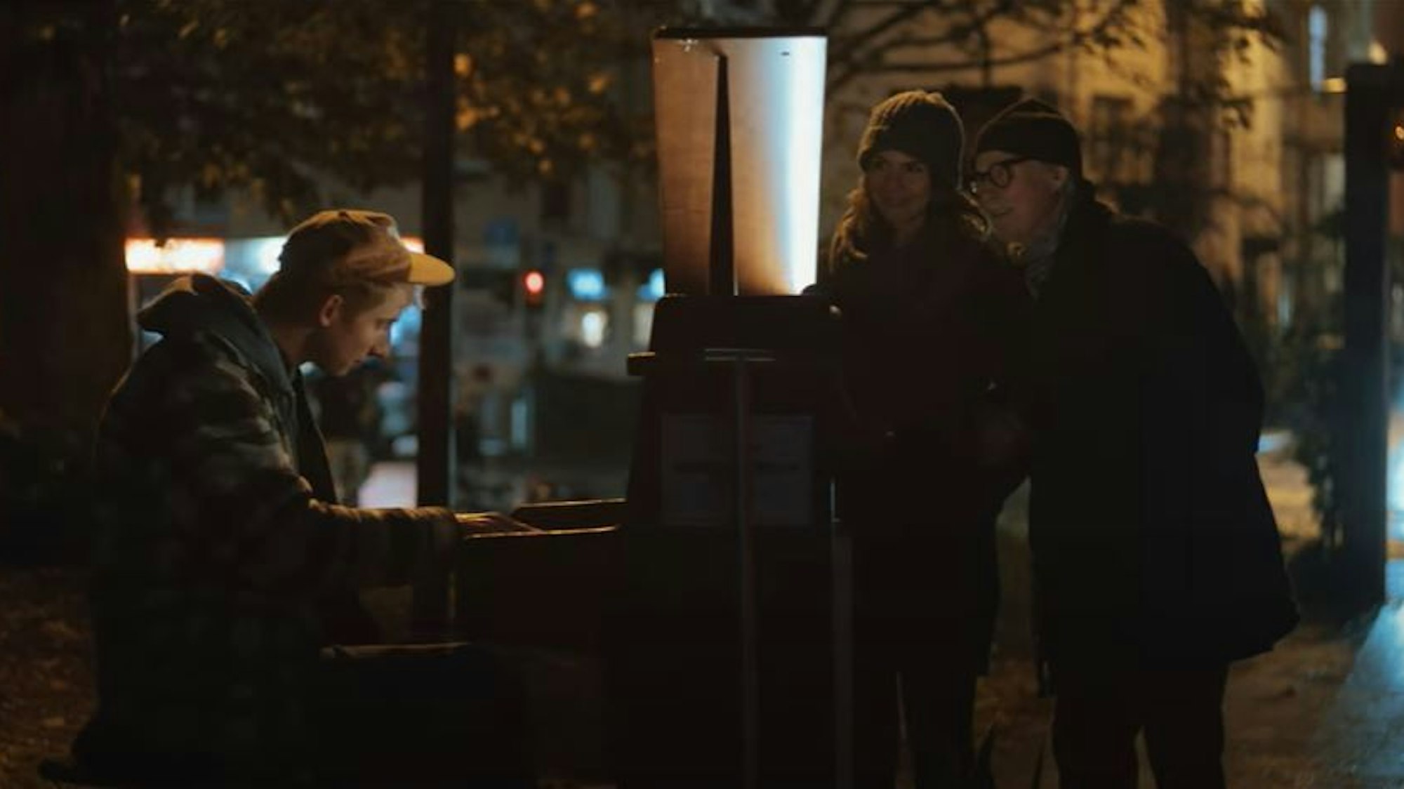 Zwei Menschen stehen vor einem Klavierspieler und hören seiner Musik zu.