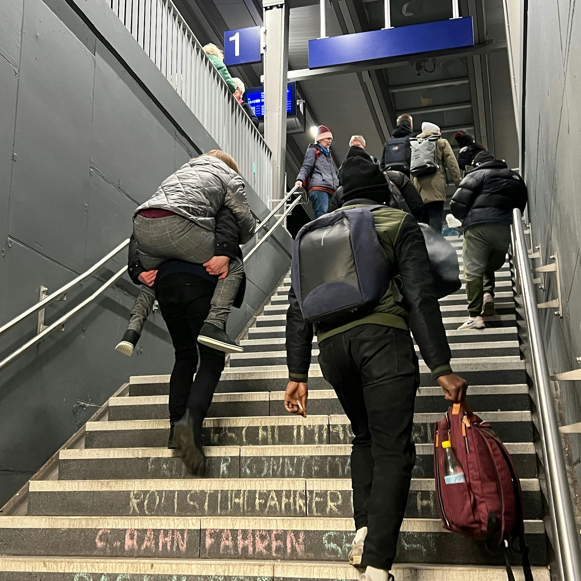 Ein Mann trägt eine gehbehinderte Frau am Bahnhof Leverkusen Mitte die Treppe zum Bahnsteig hinauf.
