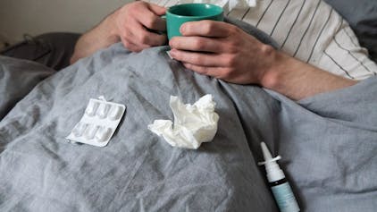 Ein Mann liegt mit Taschentüchern, Teetasse, Nasenspray und Tabletten im Bett.