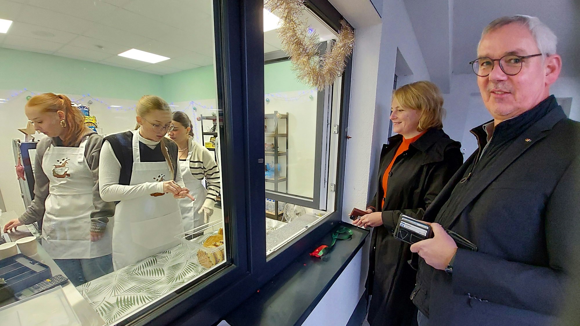 Hinter einer Glasscheibe arbeiten Schülerinnen in einer Küche; am Verkaufsfenster stehen eine blonde Frau und ein Mann.