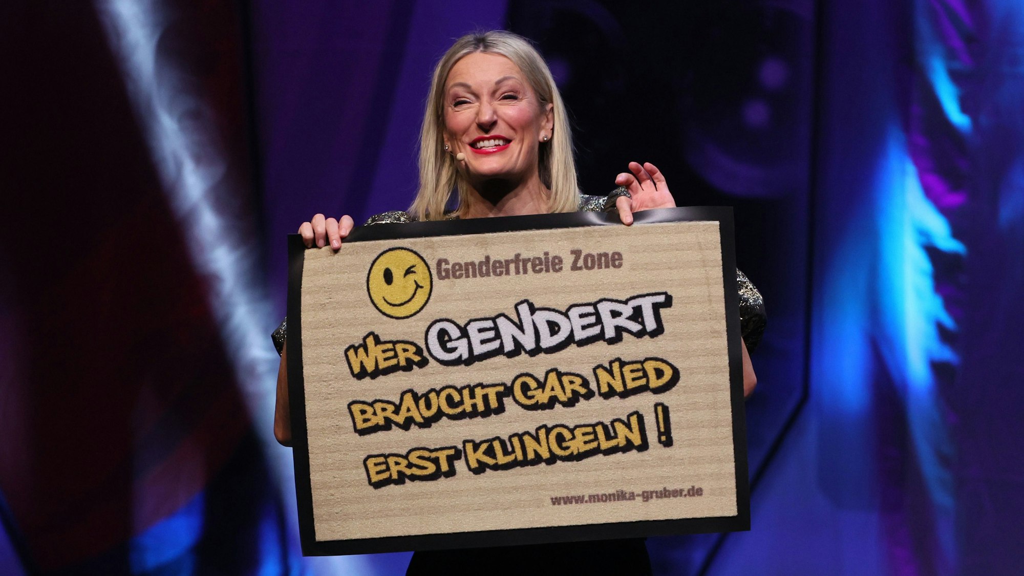 Monika Gruber steht in der Lanxess-Arena und zeigt eine Fußmatte, auf der steht: „Wer gendert, braucht gar ned erst klingeln!“