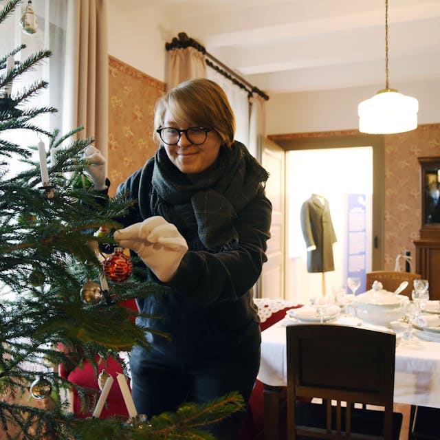 Eine Frau schmückt einen Weihnachtsbaum. Sie trägt weiße Wollhandschuhe, um die Glaskugeln nicht zu beschädigen.&nbsp;