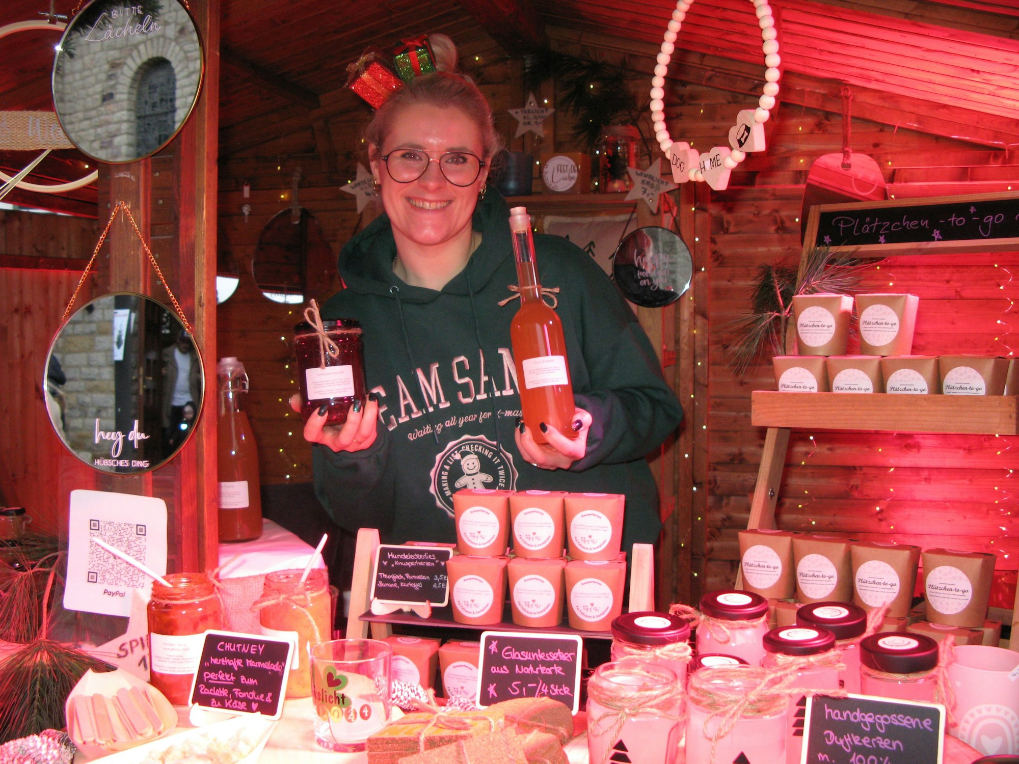 Eine Frau in einem Weihnachtsmarktstand. Sie hält Marmelade und selbst hergestellten Likör in der Hand. Im Haar hat sie einen Reifen mit kleinen Päckchen.