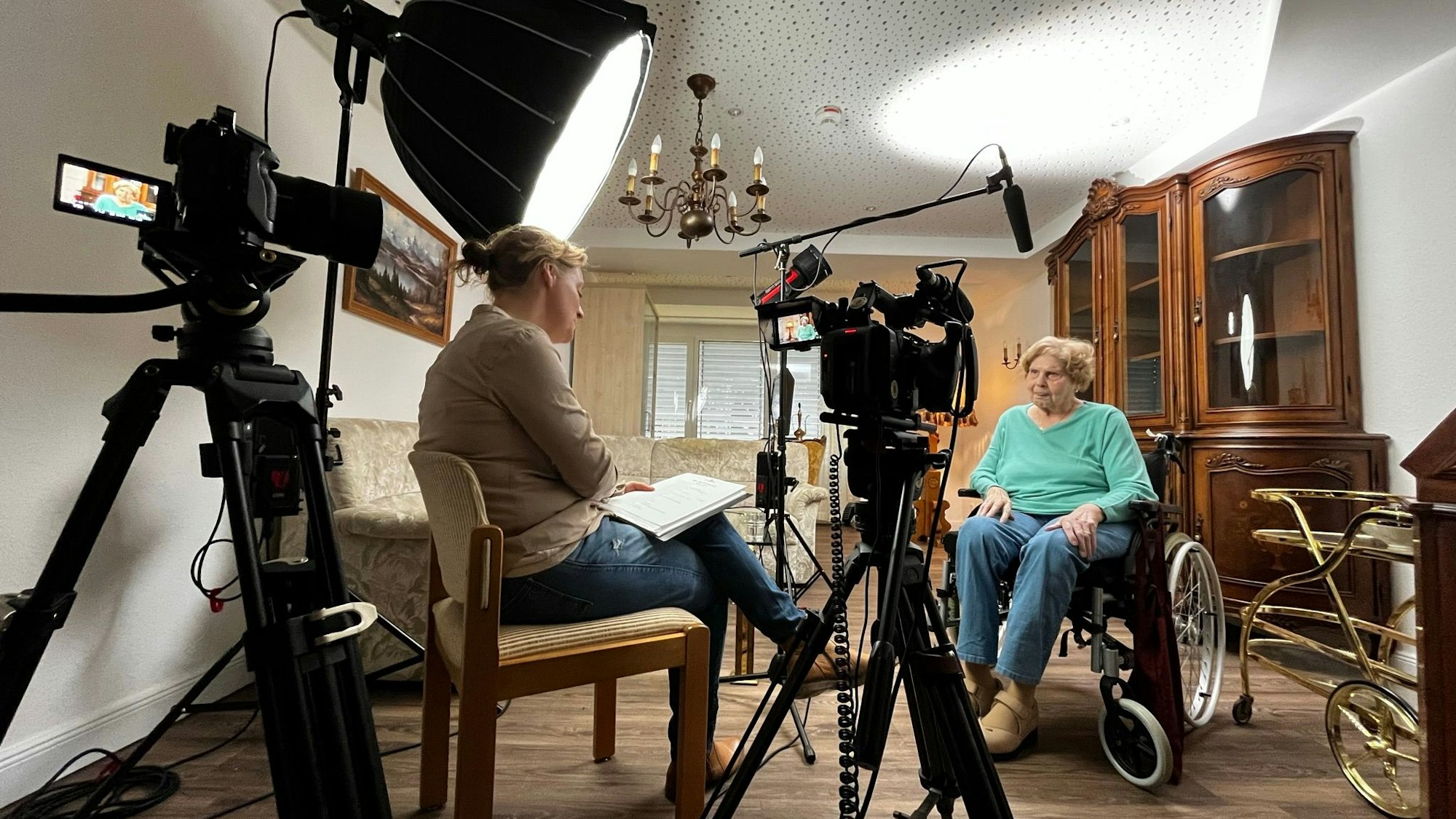 Eine mittelalte Interviewerin spricht vor laufenden Videokameras mit einer im Rollstuhl sitzenden Seniorin.