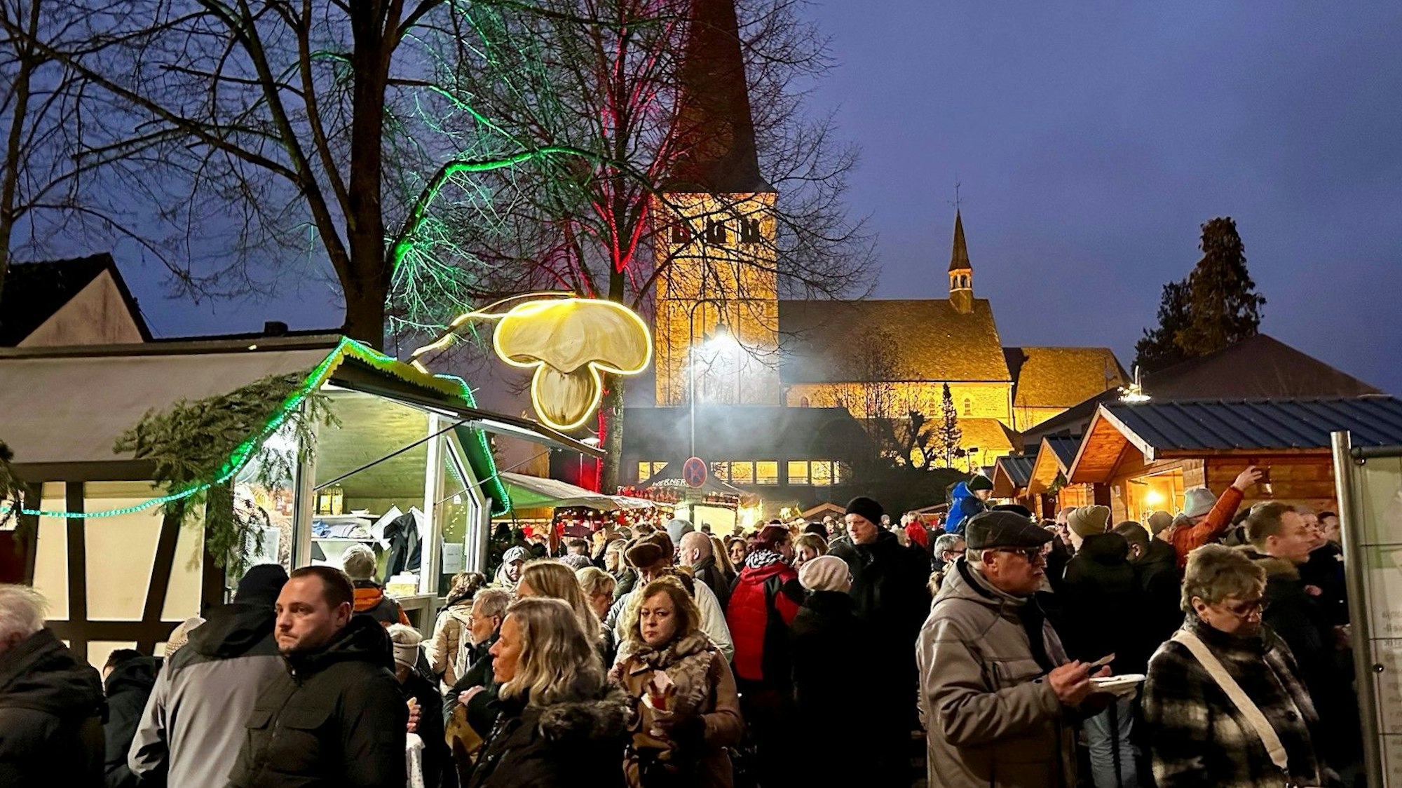 Ein Weihnachtsmarkt in der Dämmerung. Viele Menschen drängen sich in der Budengasse um beleuchtete Weihnachtshäuschen. Im Hintergrund steht eine beleuchtete Kirche.