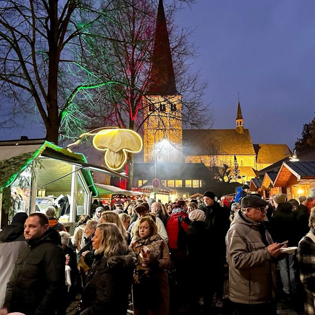 Ein Weihnachtsmarkt in der Dämmerung. Viele Menschen drängen sich in der Budengasse um beleuchtete Weihnachtshäuschen. Im Hintergrund steht eine beleuchtete Kirche.