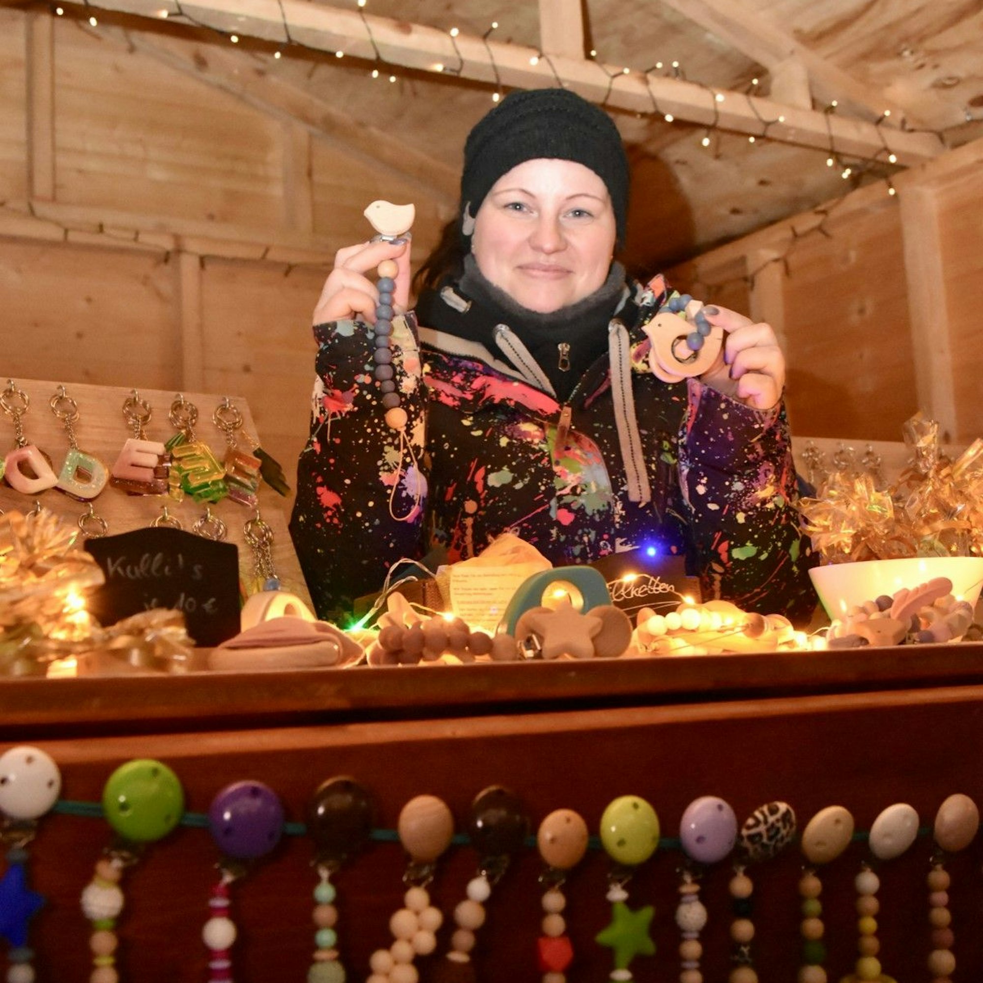 Eine Frau an ihrem Weihnachtsmarktstand. Sie zeigt Schnullerketten, die auch im Vordergrund hängen.