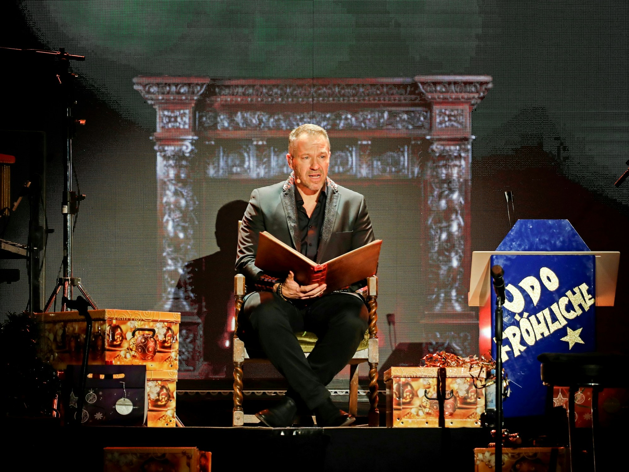 Ein Mann sitzt auf einer Bühne auf einem Stuhl und liest aus einem Buch.