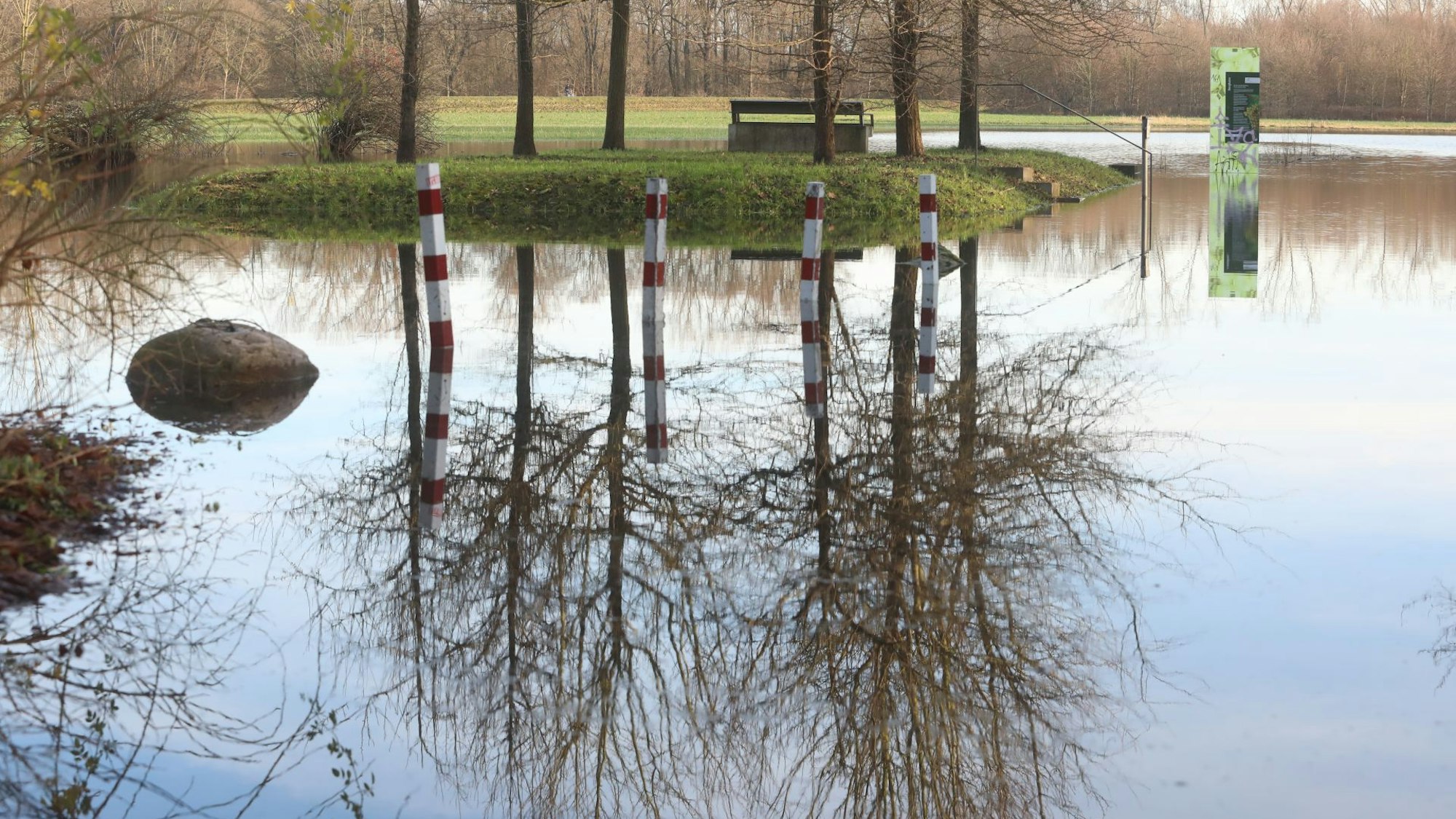 im Wasser auf einem überfluteten Weg spiegeln sich Bäume und rot-weiße Pfosten.