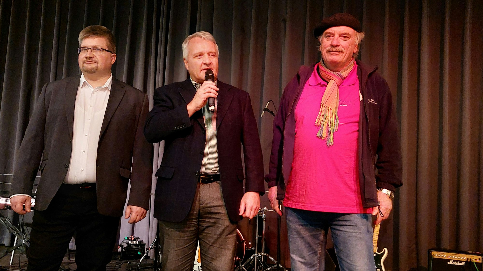 Drei Männer auf einer Bühne; der Mann in der Mitte spricht in ein Mikrofon.