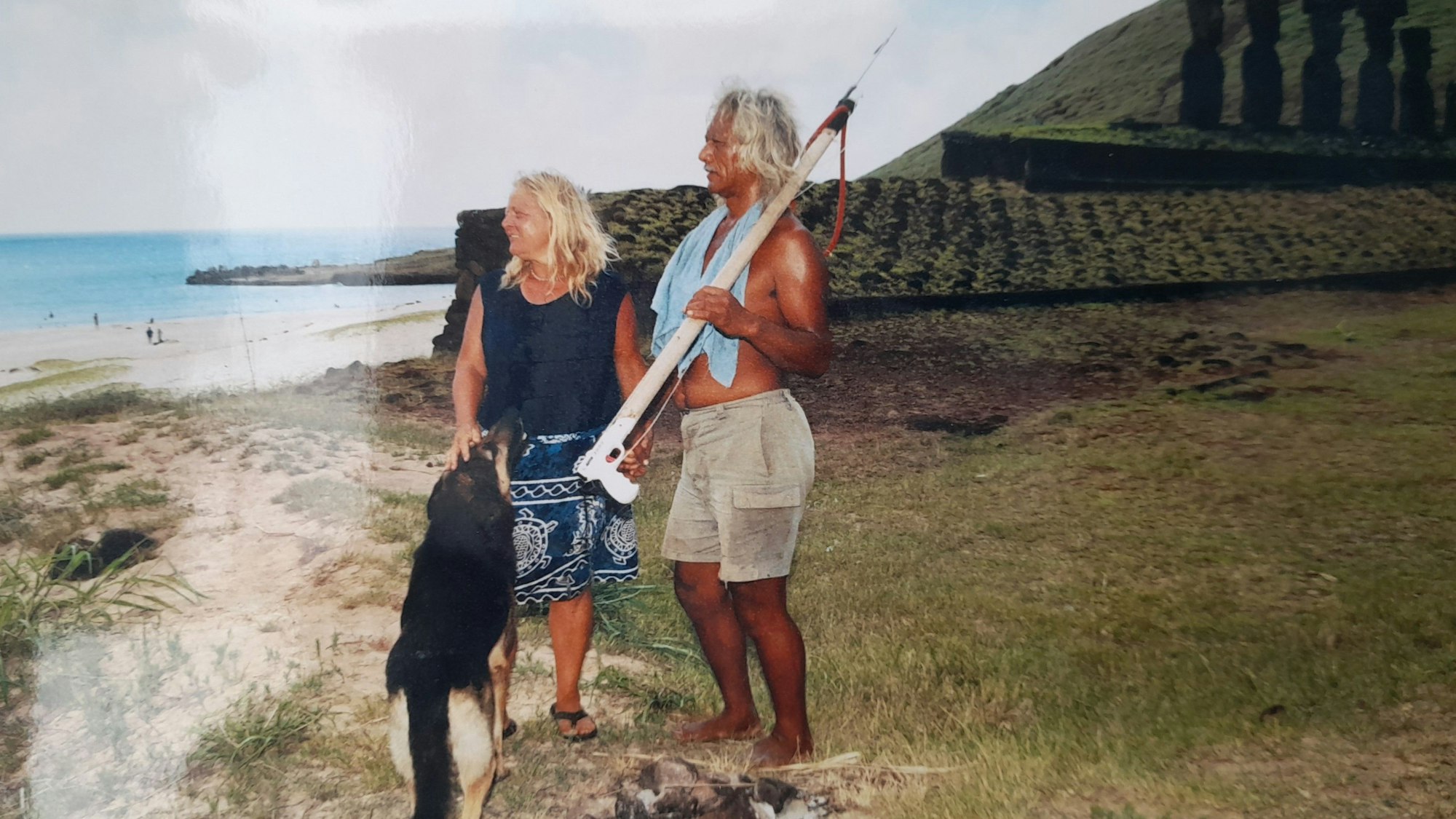 Stephanie Pauly steht mit ihrem Partner Karlo und Hund vor einer Gebetsstätte. Im Hintergrund der Strand. Karlo hat eine Harpune in der Hand.