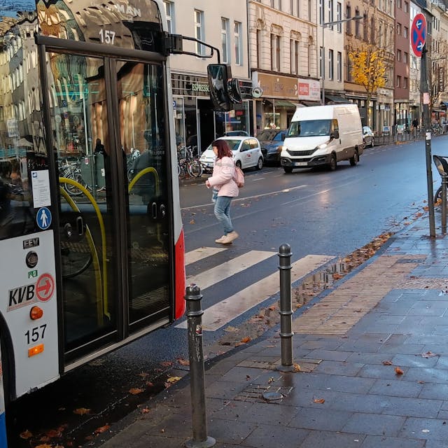 Eine Frau und eine Radfahrerin queren die Bonner Straße auf einem Zebrastreifen, vor dem ein Bus wartet.