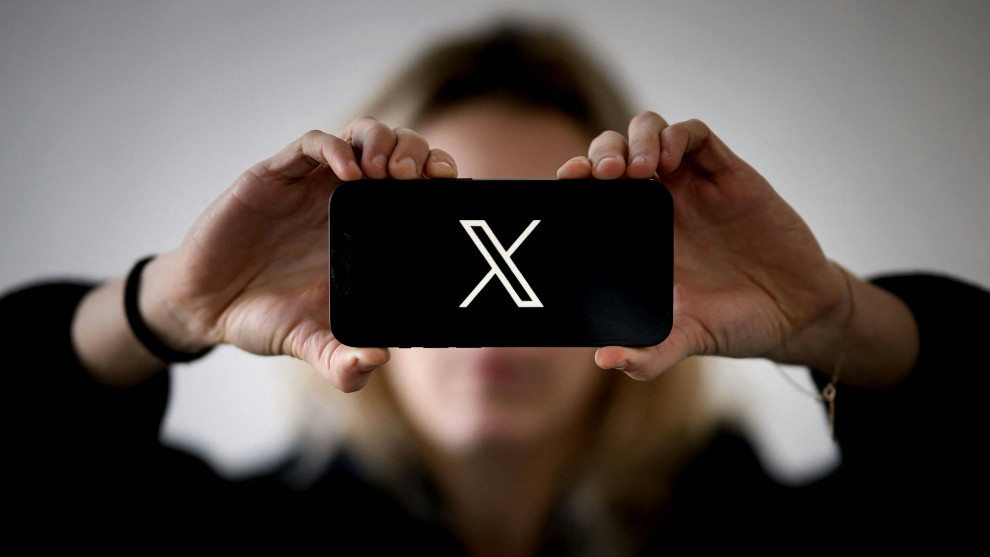 Eine Frau hält ein Smartphone in den Händen, auf dem das Logo von X zu sehen ist. (Symbolbild)