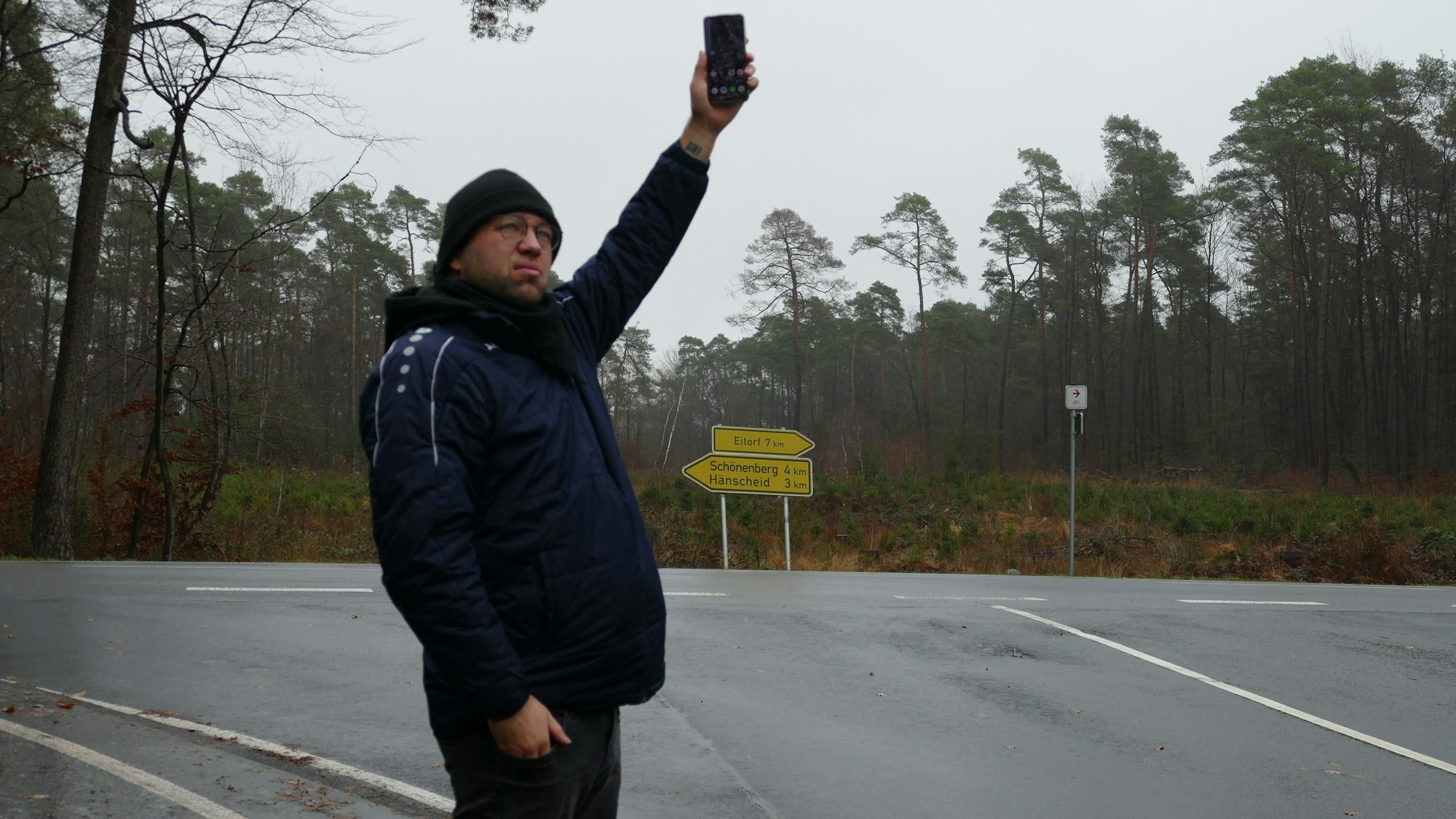 Ein junger Mann steht an einer Kreuzung und hält sein Handy hoch.