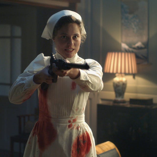 Krankenschwester Johanna Gabathuler (Dominique Devenport) hält eine Waffe in Richtung Kamera. Ihre weiße Kleidung ist voller Blutflecken.