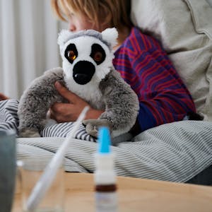 Ein Fieberthermometer, Nasenspray und eine Tasse Tee stehen vor einem Bett, in dem ein Kind mit Kuscheltier sitzt und auf ein Tablet schaut.&nbsp;