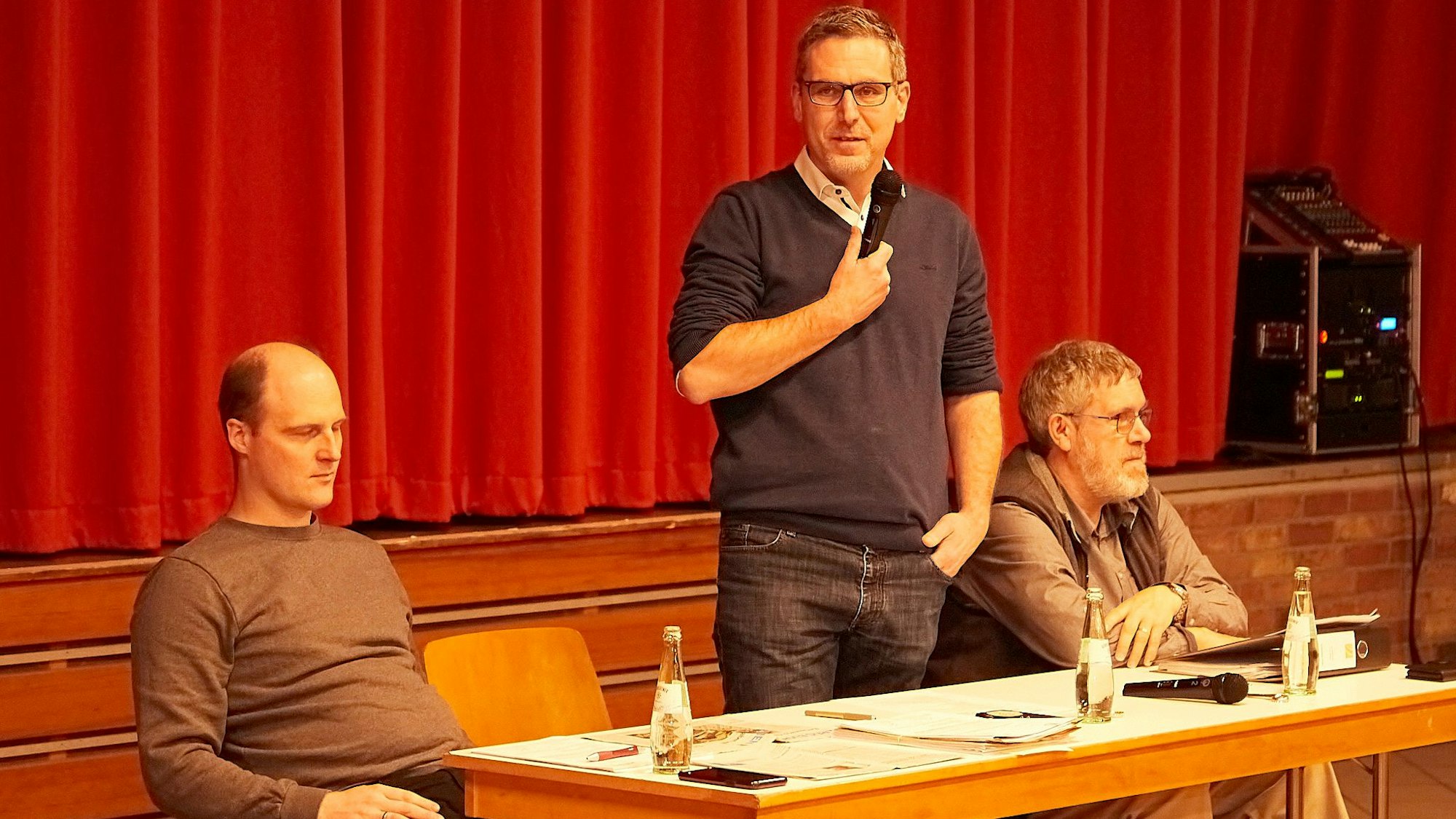 An einem Tisch vor einem roten Vorhang sitzen Simon Jung und Rainer Breinig. Nettersheims Bürgermeister Norbert Crump steht in der Mitte und hält ein Mikrofon in der Hand.