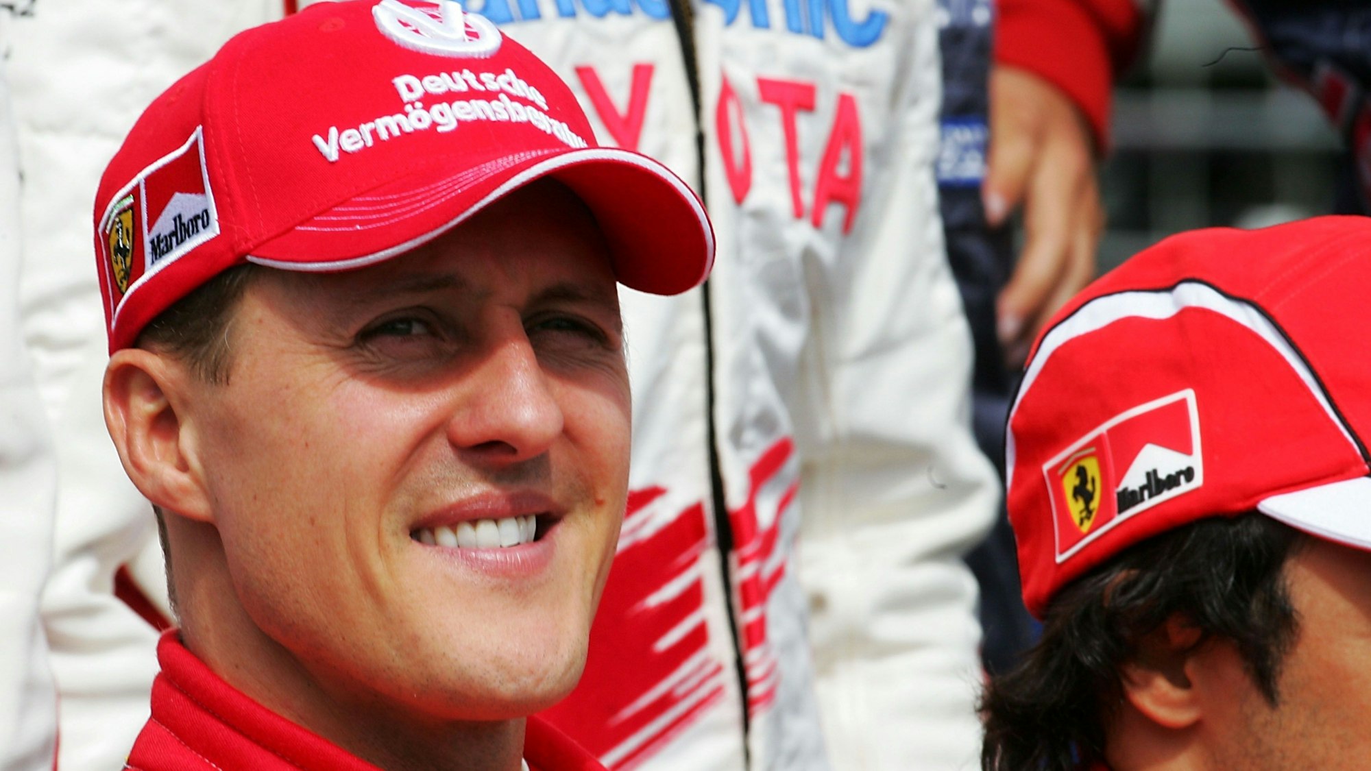 Michael Schumacher lächelt. Er trägt seinen roten Rennanzug und eine rote Kappe aus seiner Zeit bei Ferrari.
