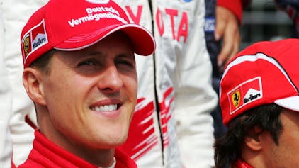 Michael Schumacher lächelt. Er trägt seinen roten Rennanzug und eine rote Kappe aus seiner Zeit bei Ferrari.&nbsp;