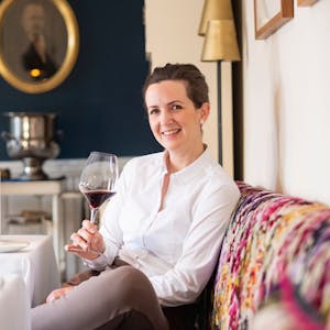 Katharina Röder sitzt in weißem Hemd mit einem Glas Wein in der Hand auf einer bunt gepolsterten Sitzbank an einem weiß gedeckten Tisch.