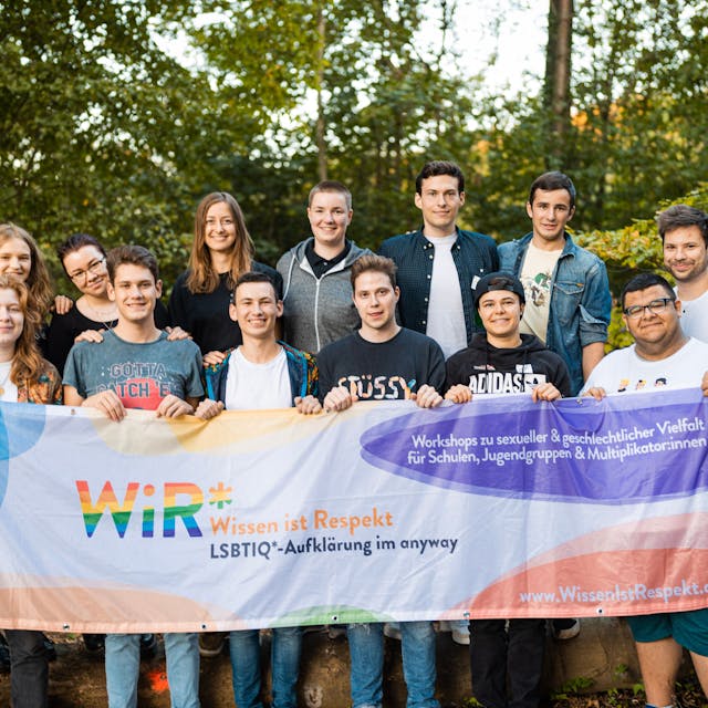 13 jugendliche Multiplikatorinnen und Multiplikatoren des „WiR*"-Projekts, das das „anyway “Kölner Jugendlichen anbietet, halten eine Fahne hoch, auf der „Wissen ist Respekt“ geschrieben steht.