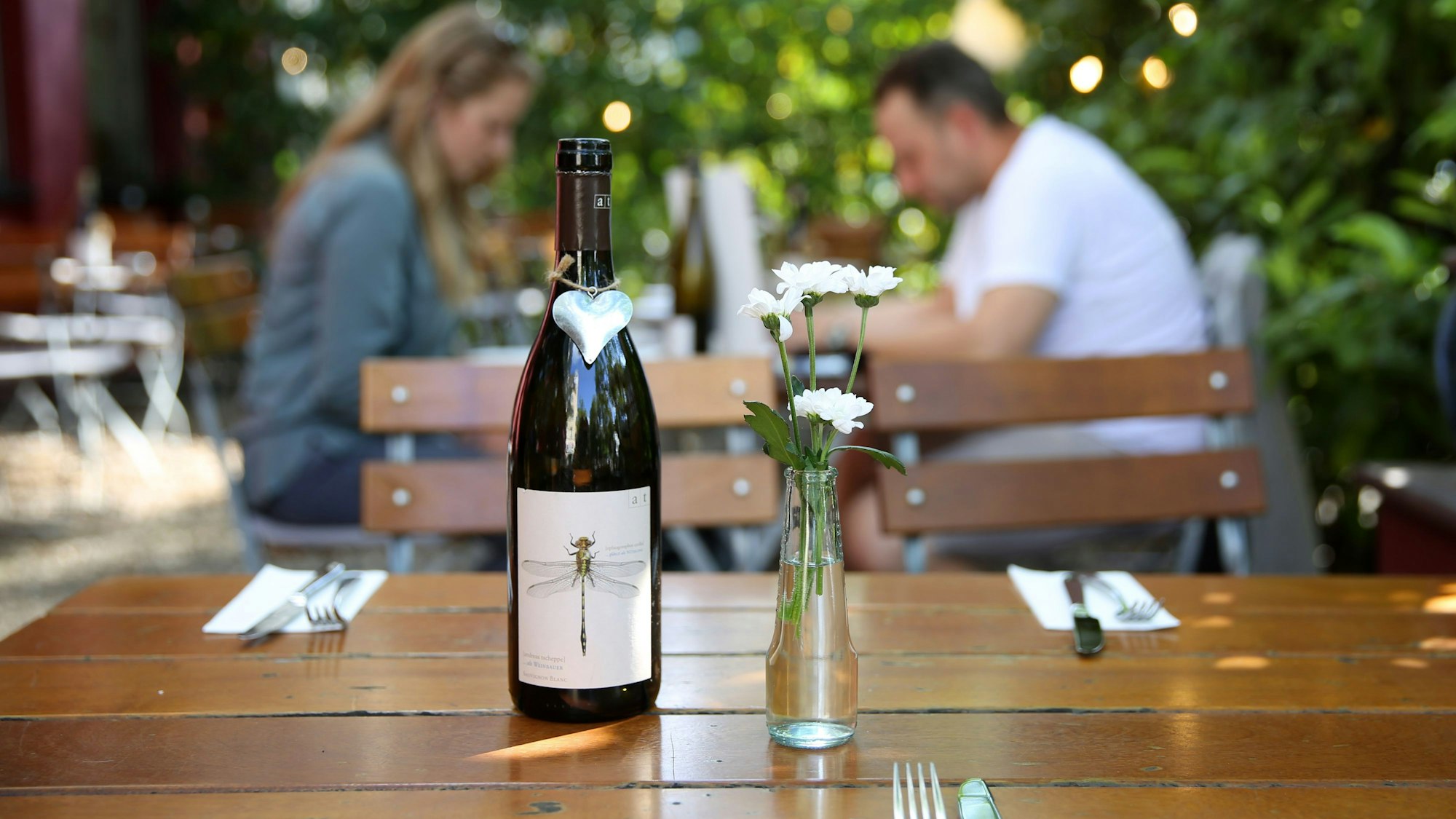 Die Außengastronomie von Essers Gasthaus: Eine Flasche mit einer Libelle auf dem Etikett steht neben einer kleinen Blumenvase auf einem Holztisch, im unschafren Hintergrund essen Gäste.