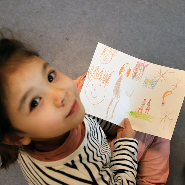 Ein Mädchen hält einen gemalten Wunschzettel in der Hand und blickt in die Kamera.&nbsp;