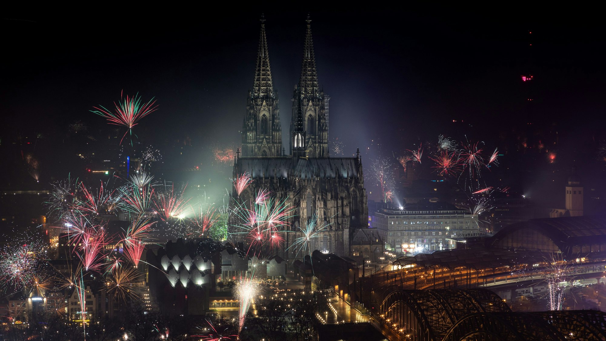Feuerwerk explodiert zum Jahreswechsel rund um den Kölner Dom, dieser und die Hohenzollernbrücke ist angeleuchtet.