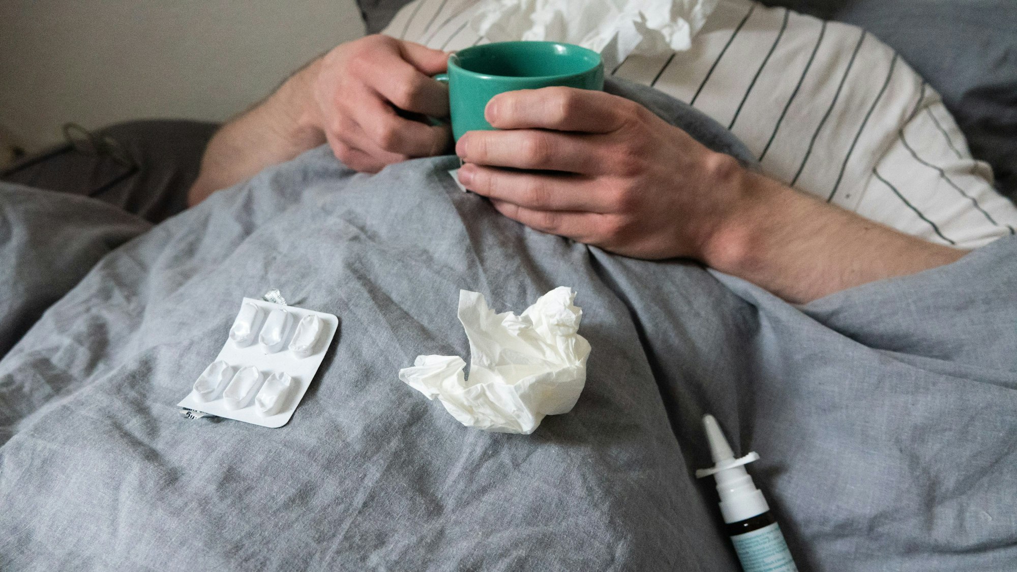 Ein Mann liegt mit Taschentüchern, Teetasse, Nasenspray und Tabletten im Bett (gestellte Szene): Mehr als jeder Zehnte ist noch oder war gerade betroffen von einer Infektion.