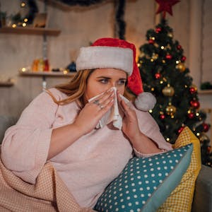 Eine Frau mit Weihnachtsmütze auf dem Kopf liegt auf dem Sofa vor einem Tannenbaum und schnäuzt sich die Nase.