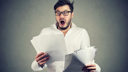 Young shocked guy in glasses looking at papers bank documents with list of bills and fees and feeling stressed on gray background