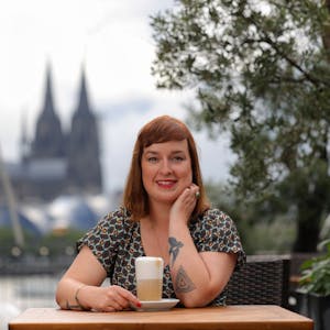 Julia Floß steht mit einem Latte macchiato an einem Stehtisch mit dem Kölner Dom im Hintergrund.