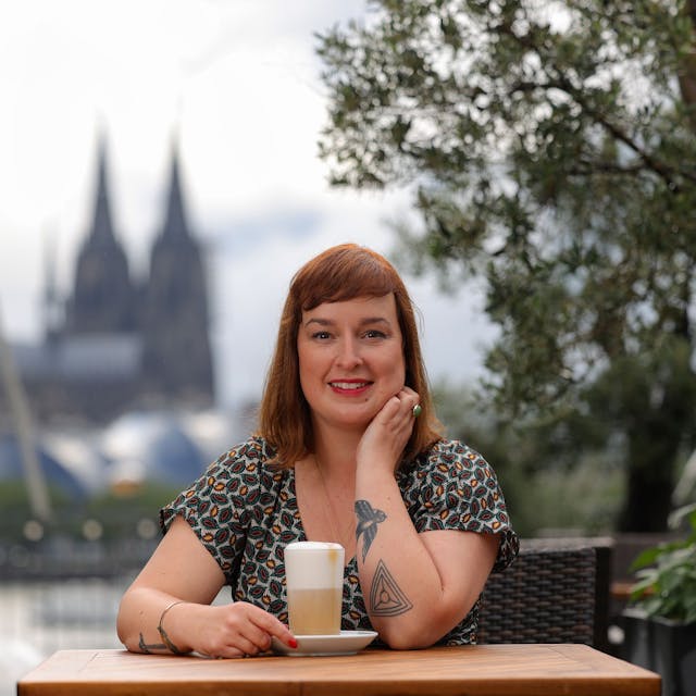 Julia Floß steht mit einem Latte macchiato an einem Stehtisch mit dem Kölner Dom im Hintergrund.