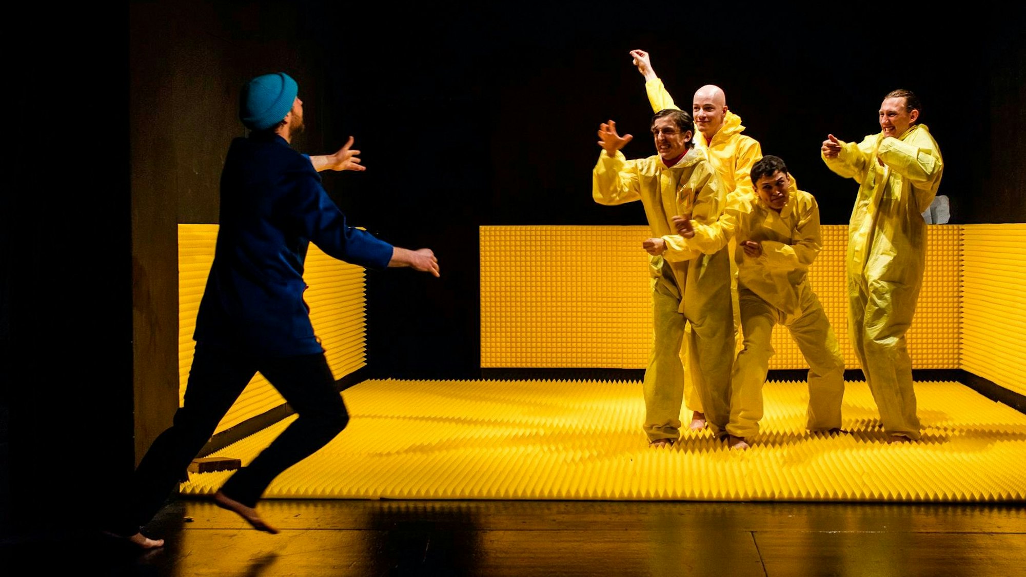 Vier Schauspieler in gelben Anzügen stehen auf einer gelben Gummimatte. Ein Schauspieler in Blau läuft auf sie zu.
