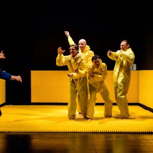 Vier Schauspieler in gelben Anzügen stehen auf einer gelben Gummimatte. Ein Schauspieler in Blau läuft auf sie zu.