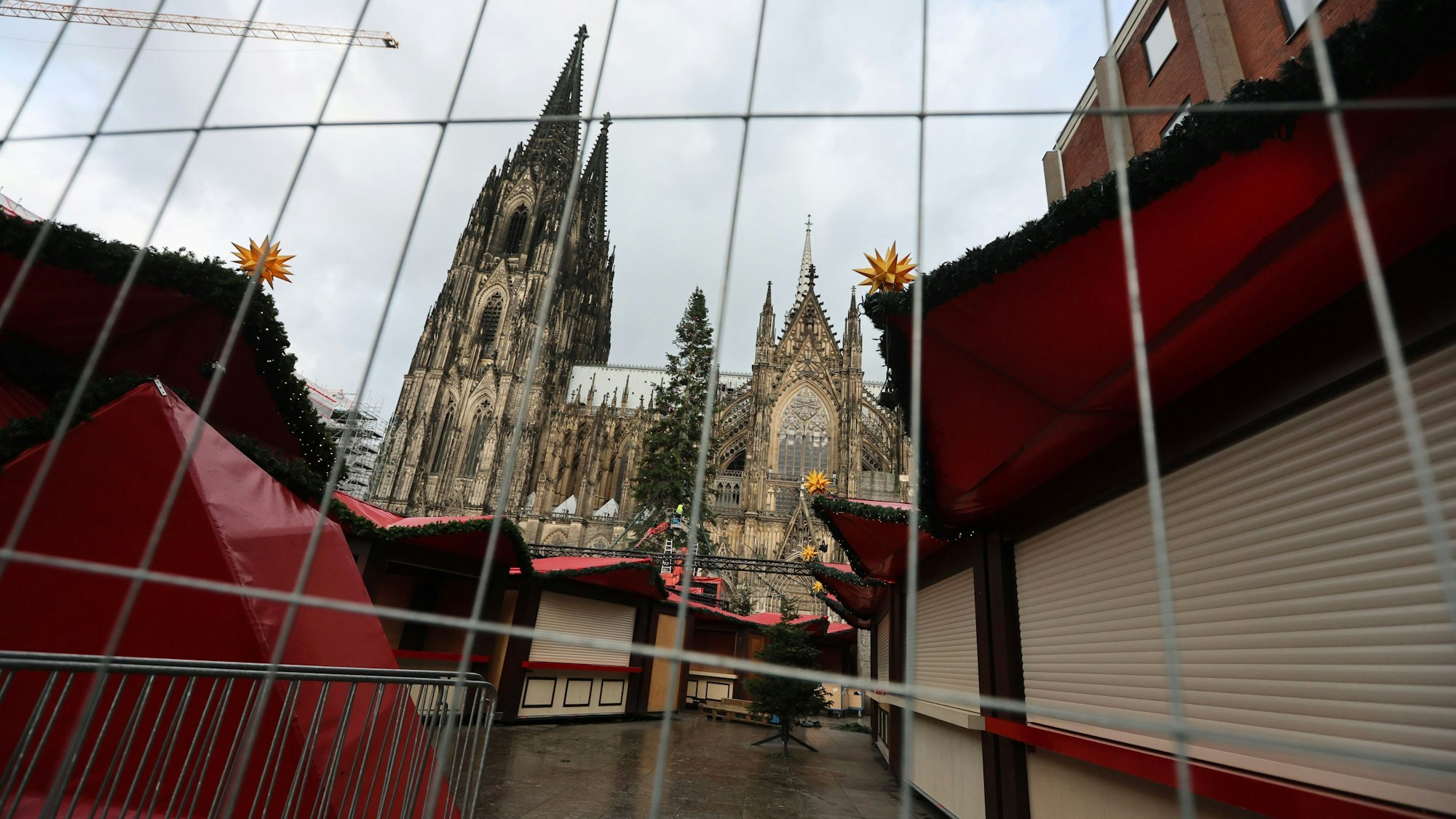 Durch einen Bauzaun sind leerstehende Buden des Weihnachtsmarkts am Kölner Dom fotografiert, im Hintergrund erhebt sich die Kathedrale.