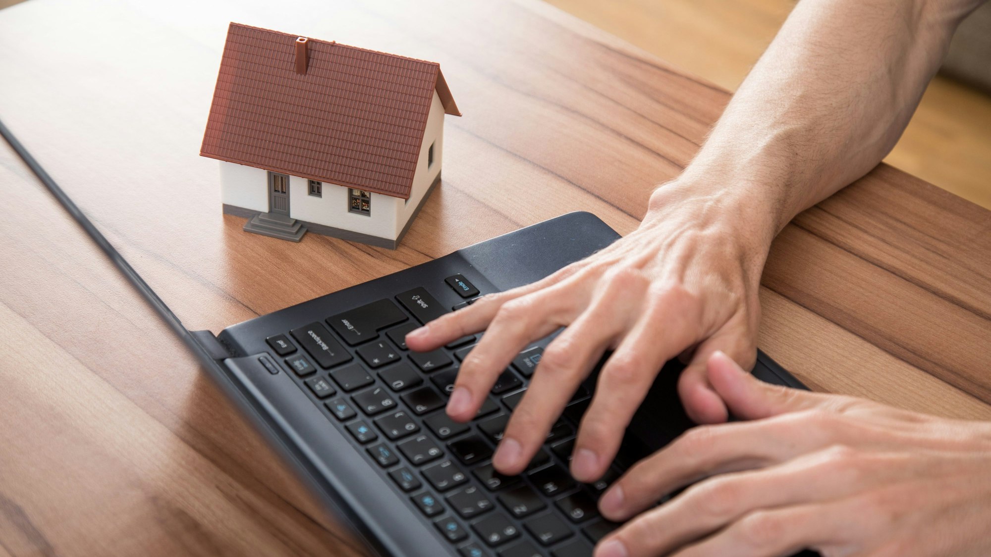 Hände tippen auf eine Tastatur, daneben steht ein Miniaturmodell eines Hauses auf dem Tisch.