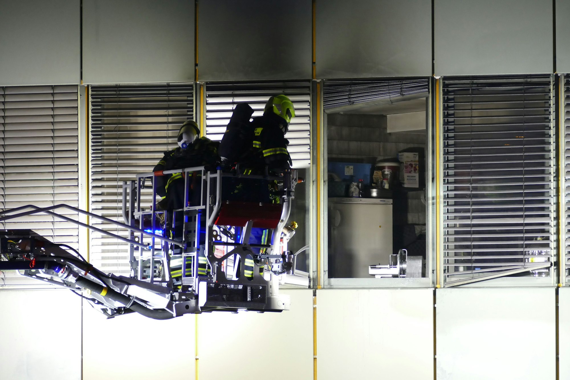 Feuerwehrleute stehen im Korb einer Drehleiter vor einer Fassade mit geöffneten Fenstern.