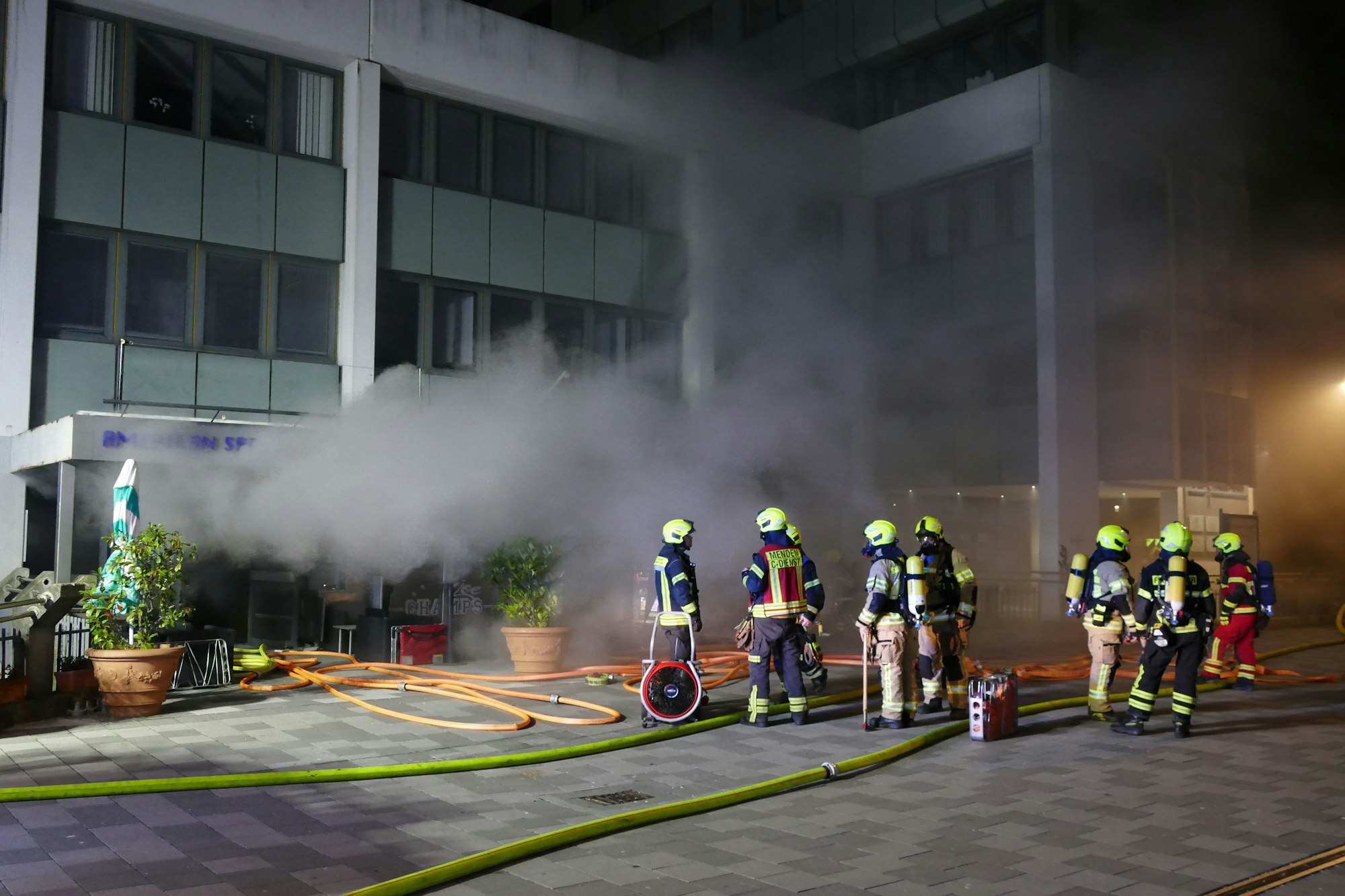 Rauch kommt aus einem Gebäudeeingang, davor stehen Feuerwehrleute.