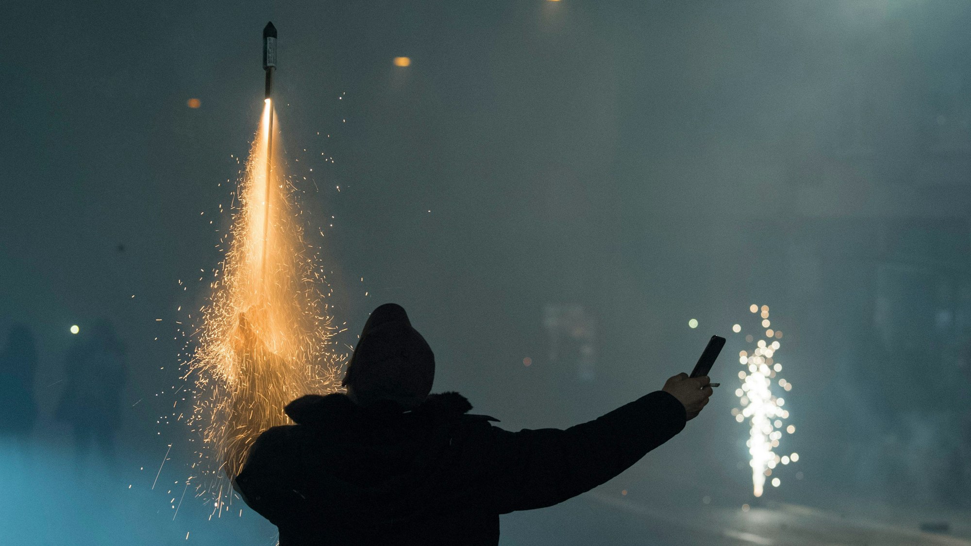 Eine Person hält auf einer Straße in der einen Hand eine zündende Rakete, in der anderen einen Böller, im Hintergrund Rauchschwaden von Feuerwerk.