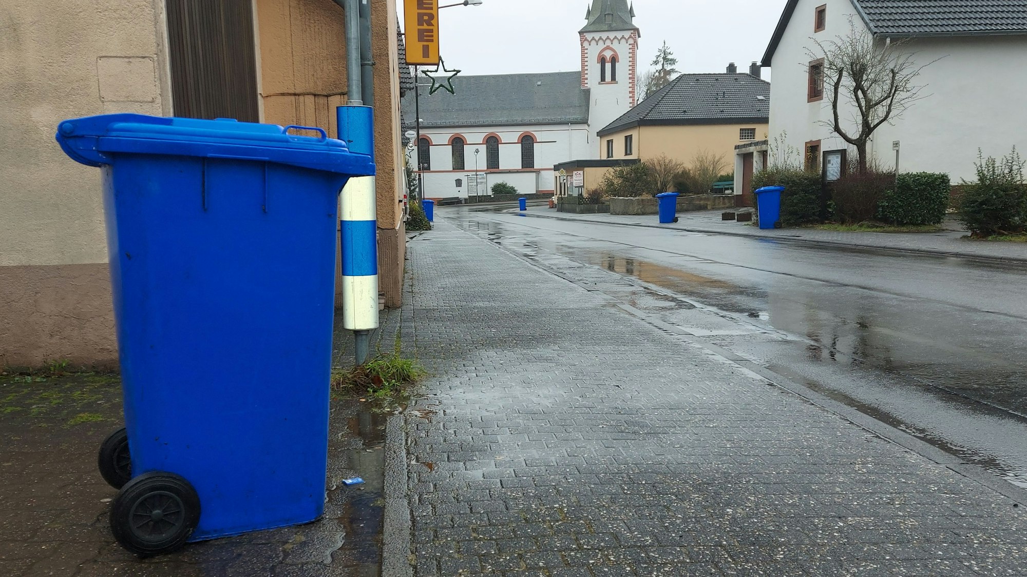 Eine blaue Altpapier-Tonne steht am Straßenrand. Im Hintergrund ist eine Kirche zu sehen.