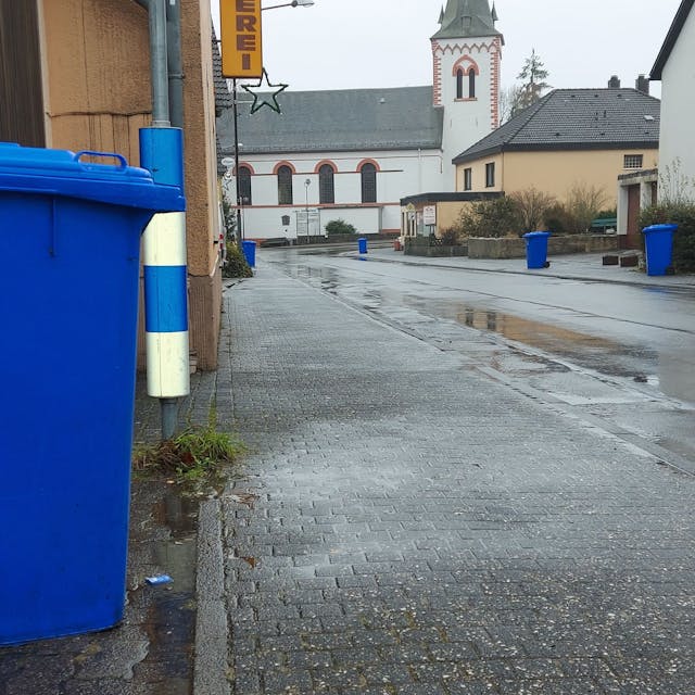Eine blaue Altpapier-Tonne steht am Straßenrand. Im Hintergrund ist eine Kirche zu sehen.