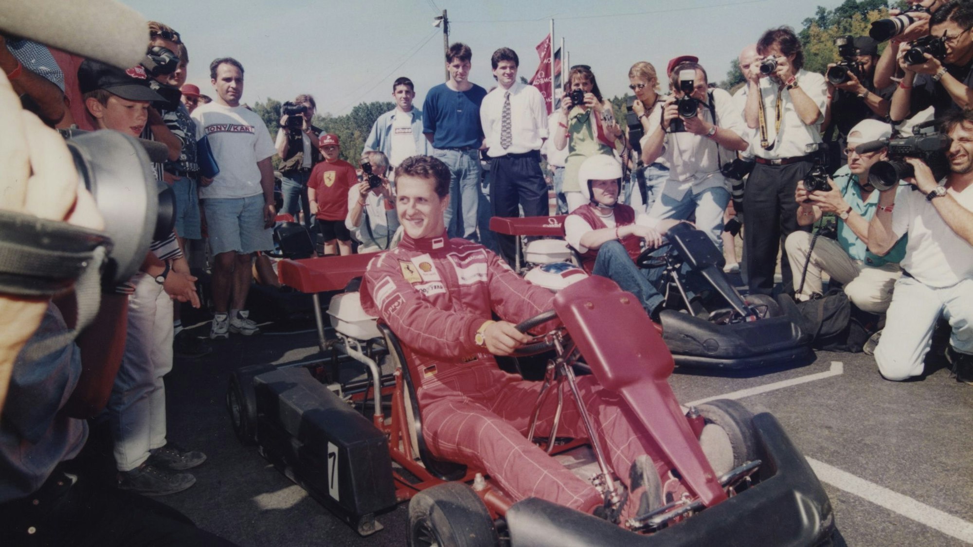 Michael Schumacher sitzt in einem roten Ferrari-Overall auf einem Kart umgeben von Fotografen.
