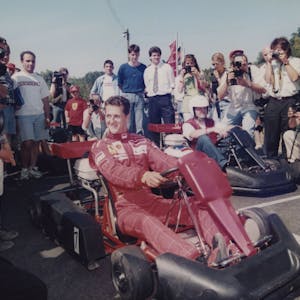 Michael Schumacher sitzt in einem roten Ferrari-Overall auf einem Kart umgeben von Fotografen.