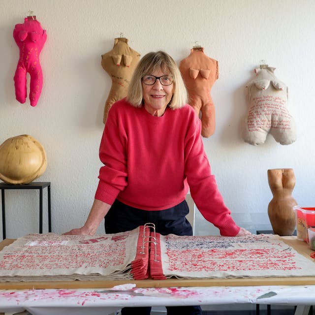 Frau in rotem Pulli in ihrem Atelier vor einem großen, vollgeschriebenen Buch. Die Schrift ist gestickt.