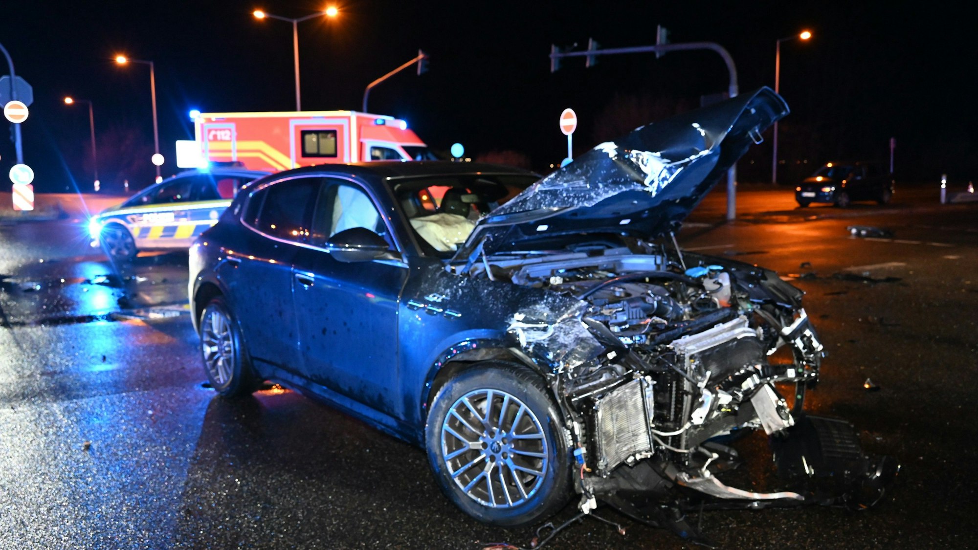 Ein schwer beschädigter Maserati steht auf einer Straßenkreuzung. Im Hintergrund sind ein Polizeiwagen und ein Rettungswagen zu sehen