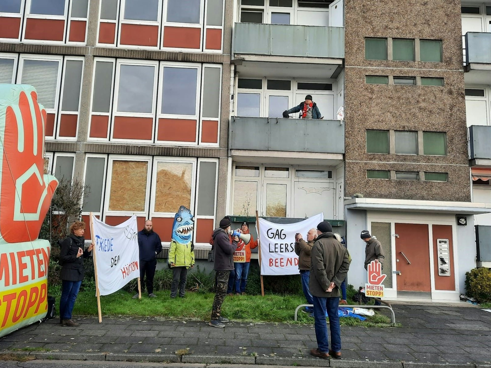 Demo-Teilnehmende stehen mit Plakaten vor einem mehrstöckigen Haus, auf einem Balkon in der ersten Etage steht Aktivist Rainer Kippe.
