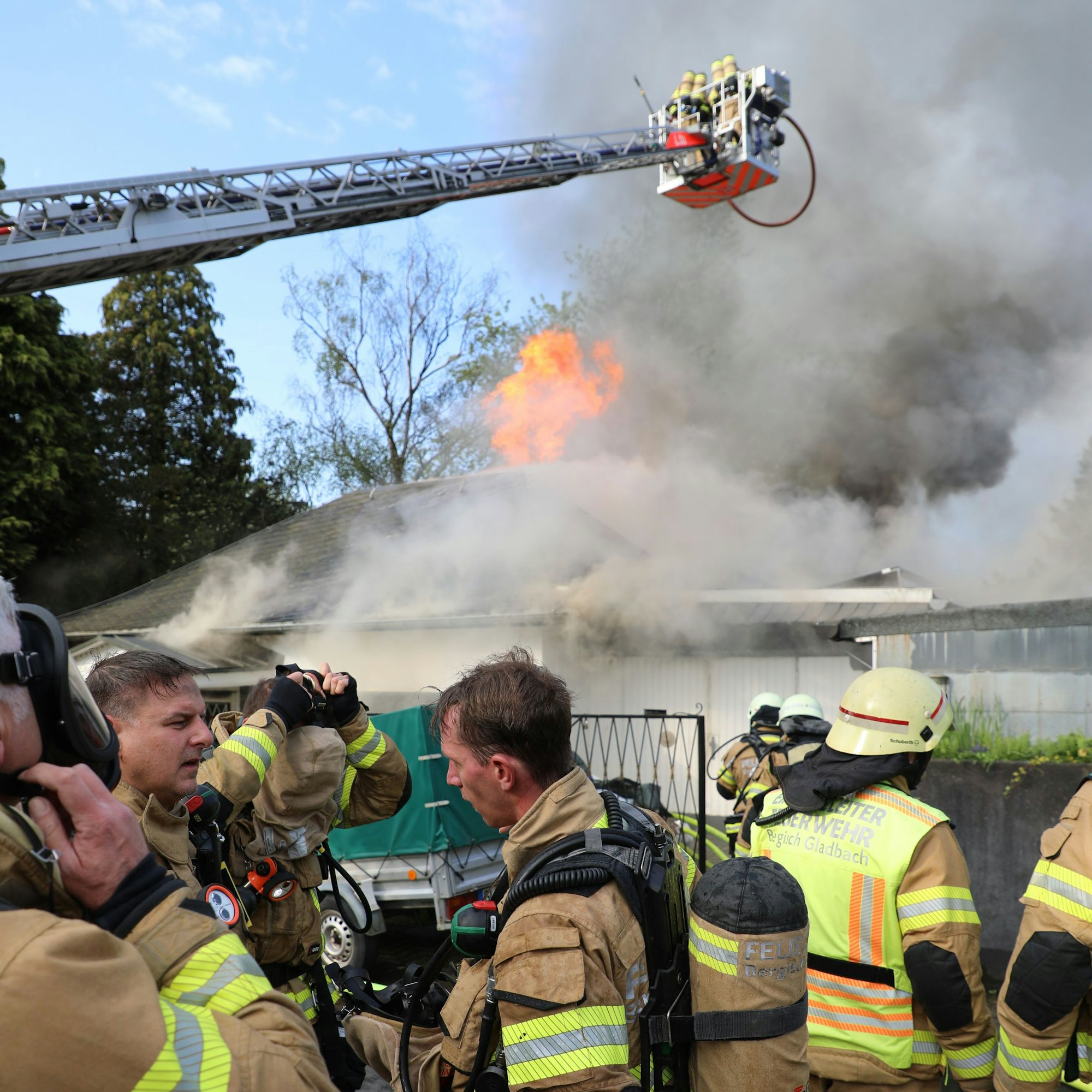Feuerwehrleute stehen vor einem brennenden Haus, das auch von einer Drehleiter aus gelöscht wird.