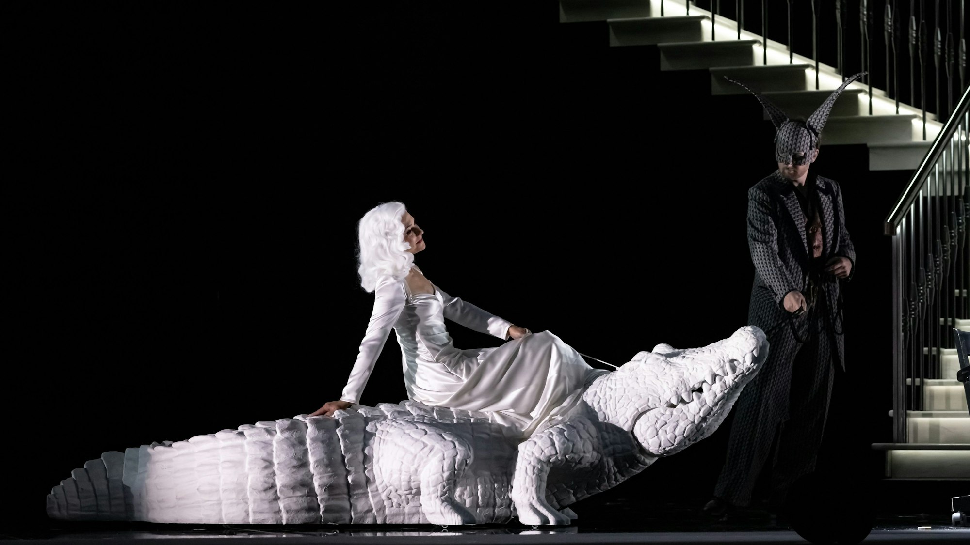 Eine ganz in Weiß gekleidete Frau reitet ein weißes Krokodil. Ein Mann guckt erstaunt.
