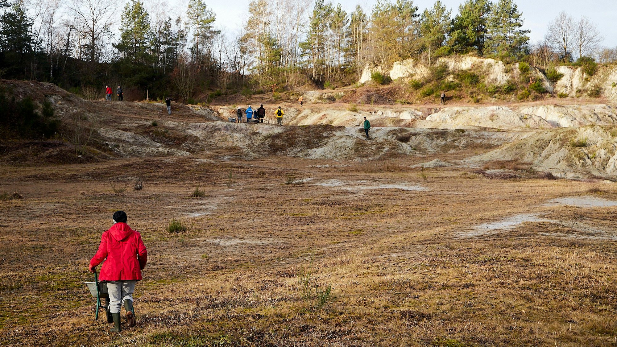 Ein Mann in roter Jacke schiebt eine Schubkarre über ein offenes Gelände einer ehemaligen Tongrube.